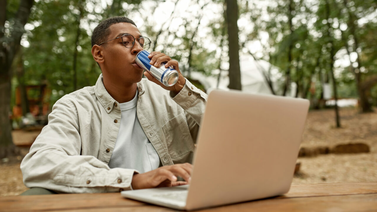 A man is working on his laptop outdoors while sipping on a branded energy drink. Coffee vs energy drinks concept